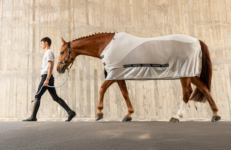 Ein Reiter führt ein Pferd an der Hand Das Pferd trägt eine Sommerdecke von Sunride in hellgrau