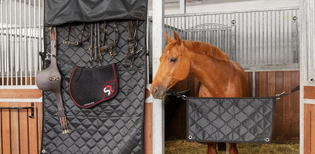 Ein schwarzer Boxenvorhang hängt an einer Pferdebox daran befestigt sind eine schwarze Schabracke mit rotem Rand und ein Reithelm rechts daneben schaut ein braunes Pferd aus seiner Box