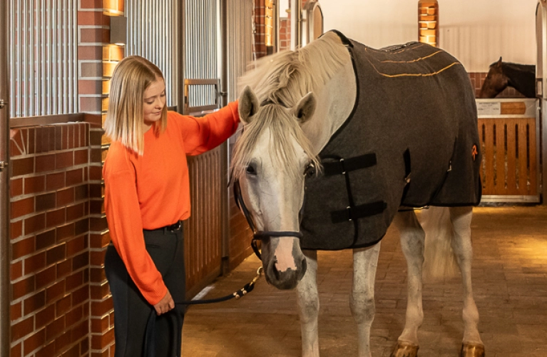 Eine Reiterin steht in der Stallgasse neben einem Pferd das eine Heizdecke trägt und streichelt es